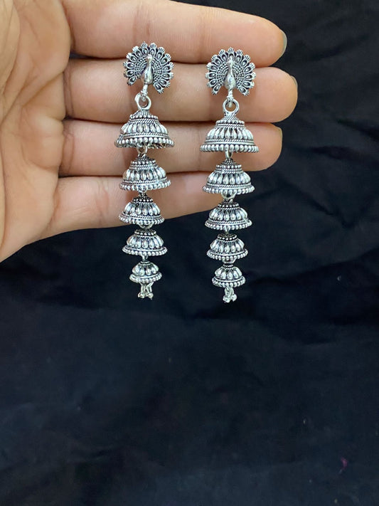 Hand holding a pair of silver jhumka earrings with intricate designs and multiple tiers.