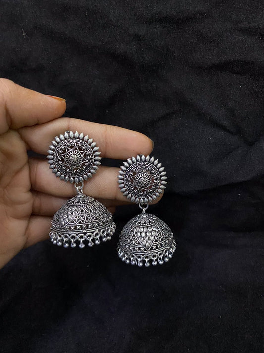 A hand holding a pair of ornate silver jhumka earrings with intricate designs.