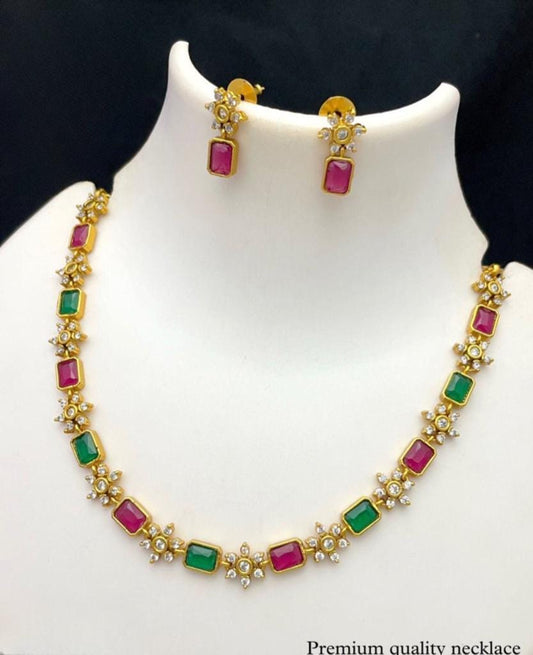 Gold necklace with alternating green and pink rectangular gemstones, adorned with small white stones, accompanied by matching earrings.